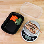Open black meal prep container with clear lid nearby and food scale showing grilled skewers on a wood surface.