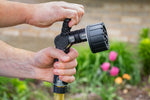 Hands holding an Orbit 6-pattern lever-control hose nozzle in a garden setting with blurred flowers
