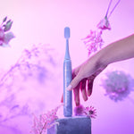 Lavender Ordo sonic toothbrush handle with decorative pattern held above a pedestal surrounded by soft floral props and pink-purple lighting