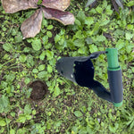 Overhead view of a black and green bulb planter resting on leafy ground beside a freshly removed soil plug
