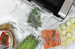 Overhead view of vacuum-sealed bags with asparagus, salmon and sliced zucchini beside a sealer