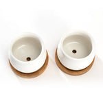 Overhead view of two white jar-shaped ceramic pots on bamboo saucers showing drainage holes at the bottom of each pot