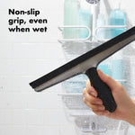 Hand holding an OXO squeegee wiping a wet glass shower door with text reading ‘Non-slip grip, even when wet’