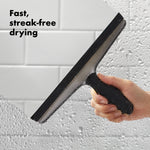 Hand using an OXO stainless squeegee on a wet tiled shower surface with text ‘Fast, streak-free drying’