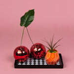 Painted orange faceted ceramic planter arranged on tray with decorative red disco balls and a leaf on pink background