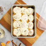 Top-down view of aluminum pan filled with frosted cinnamon rolls, with a clear plastic lid set to the side on a wooden board