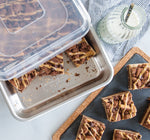 Aluminum rectangular pan partially covered by a clear plastic lid holding square dessert bars, styled with a milk bottle and napkin