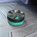 Turquoise gel air freshener with black vented lid placed on a textured car floor mat, shown from a low angle.