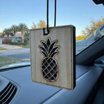 Square wooden hanging air freshener with laser-cut pineapple design suspended from a car rearview mirror