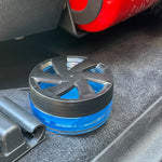 Blue gel air freshener with black vented lid placed on a car carpeted floor area near a red object
