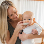 Adult smiling while using a gray silicone finger toothbrush in a baby's mouth as the baby looks forward