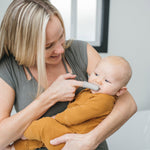 Adult holding a baby while using a gray silicone finger toothbrush gently inside the infant's mouth