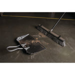Lifestyle scene of an aluminum dustpan on the floor with debris beside a push broom in a workshop setting.