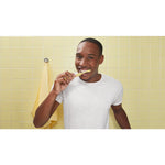 Smiling person in white shirt brushing teeth with a yellow Philips toothbrush in a yellow-tiled bathroom scene