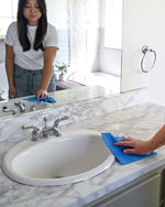 Person wiping a bathroom counter and sink with a blue textured sponge cloth in a bright bathroom