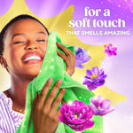 Person smiling and pressing a green towel to their face surrounded by purple flowers and text promoting a soft touch that smells amazing