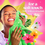 Smiling person pressing a green towel to their face surrounded by tropical flowers and text about a soft touch that smells amazing