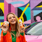 Smiling person holding a Citrus Magic Tropical Citrus spray can and solid tub in front of a colorful mural and pink car