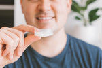 Person holding a clear thermoplastic dental night guard between fingers with a blurred smiling face in background