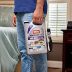 Person holding Ortho Home Defense Max gallon jug by the handle with attached Comfort Wand visible, shown standing beside a bed.