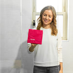 Person holding a bright pink microfibre cloth labeled GLASS while standing in front of a bathroom mirror