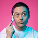 Person holding a quip toothbrush to their temple in a styled portrait with pink background and colored lighting