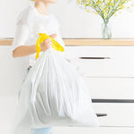 Person holding a filled white drawstring trash bag tied with a yellow drawstring in a bright kitchen setting