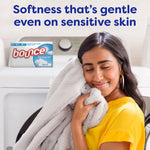 Person smiling and hugging a soft gray blanket in front of a dryer with a box of Bounce Free & Gentle visible and a softness claim