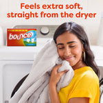 Smiling person pressing a soft blanket to their cheek in front of a washer, small Bounce box visible and softness claim