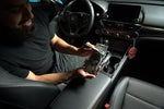 Person seated in a car holding a packaged Chemical Guys Black Frost hanging air freshener while another identical air freshener dangles from the mirror.
