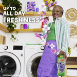 Smiling person standing in a laundry room holding a Gain scent booster bottle with washer and dryer in background and freshness text