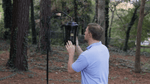 Man in a blue shirt installing or adjusting a hanging electric bug zapper in a wooded outdoor setting