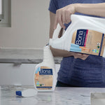 Person pouring Bona refill jug into a smaller Bona spray bottle on a kitchen countertop, spray head visible