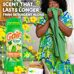 Advertising image of a person joyfully smelling green clothing with a large Gain scent booster bottle and floral graphics beside them
