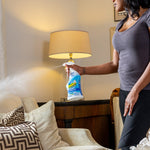 Person spraying an OdoBan disinfectant bottle toward a living room couch and lamp, visible mist in air