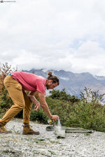 Person outdoors spraying a can of Sawyer Permethrin onto clothing laid on the ground with mountains in the background