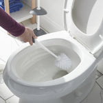 Hand holding a white toilet bowl brush with black grip cleaning the inside of a toilet bowl in a bathroom scene