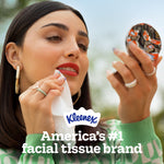 Person dabbing their face with a Kleenex tissue while looking into a compact mirror with Kleenex logo and branding text