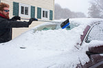 Person reaching across a snow-covered car windshield using a telescoping broom to remove heavy snow