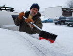 Person using a telescoping snow broom to clear snow from a vehicle roof in a snowy parking area