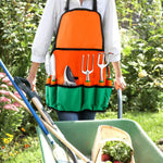 Person wearing an orange-and-green Ukoke gardening apron filled with tools, standing in a garden beside a wheelbarrow and plants.