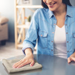 Person wiping a glossy tabletop with a folded light-gray microfiber cleaning cloth, smiling and seated at a table