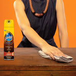 Person wiping a wooden surface with a cloth and a Pledge Moisturizing Oil spray can visible at the edge