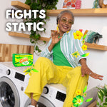 Smiling person seated on laundry machines with a small Gain dryer sheets box and decorative floral graphics
