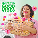 Smiling person holding a laundry basket close with floating pink and yellow flowers and text 'SNIFF THE GOOD VIBES'