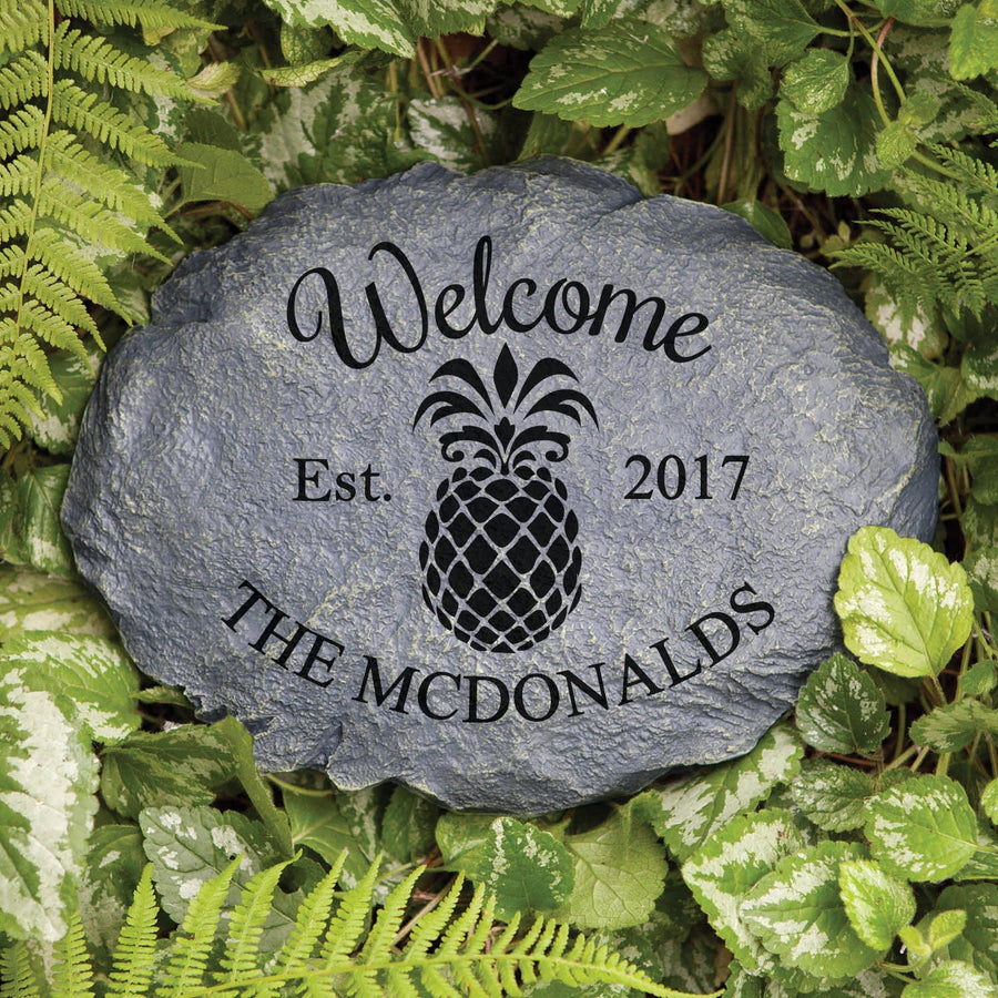 Dark gray personalized garden stone engraved “Welcome Est. 2017 THE MCDONALDS” with a pineapple motif set among green foliage.