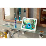 Bathroom shelf scene showing a Philips Sonicare kids toothbrush standing beside a tablet displaying a colorful animated brushing app character.