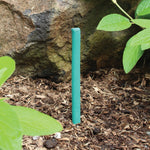 Single green mosquito-repellent stick inserted into mulch near a rock and leafy plants in a garden setting