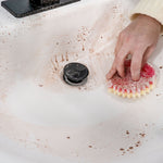 Person scrubbing a dirty white sink with a pink Scrub Mommy sponge, visible soap residue and splattered grime