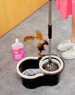 Scene with mop bucket, spilled stain and a bottle of The Pink Stuff floor cleaner staged near a mop in use